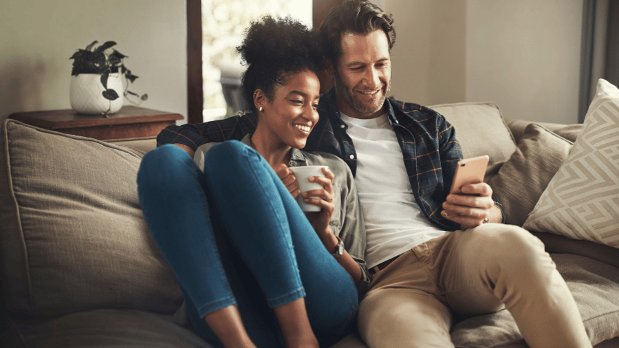 Young couple sitting on their couch at home looking at the phone together reviewing financial options