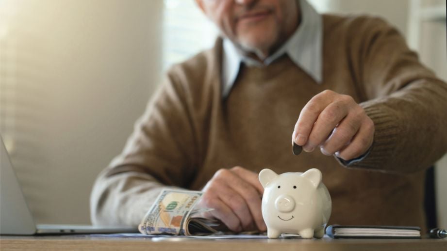 Older man in a brown sweater, adding coins into his white piggy bank. He also has a few dollar bills on the table as well, to contribute to his IRA. 