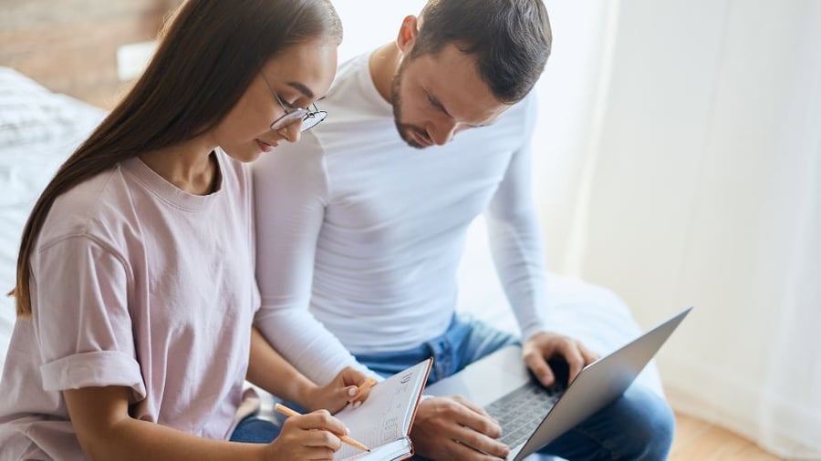 Middle aged woman and man sitting on their bed, looking at their laptop and notebook, reviewing important notes regarding their finances