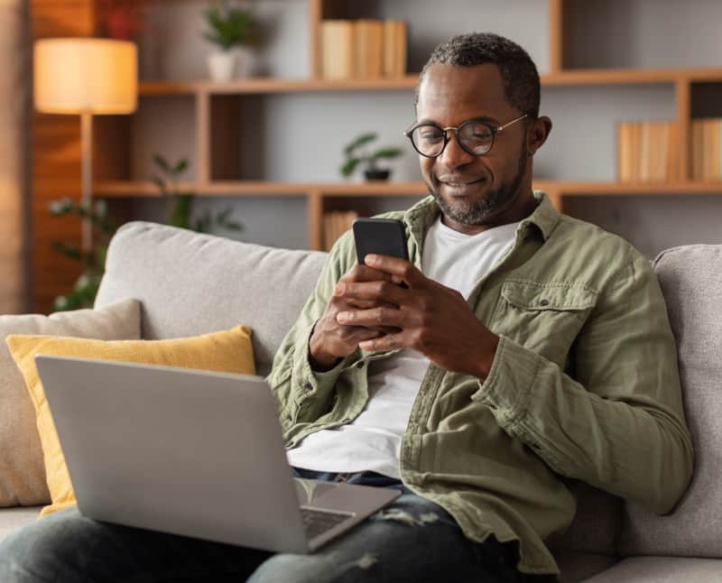 Black male in his home, wearing glasses, working on his computer to review his bank accounts. He is happy with financial freedom.