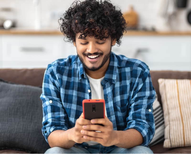 An Indian man sits on his couch in his home, completing convenient online banking. He is happily enjoying fee-free and stress-free account maintenance.